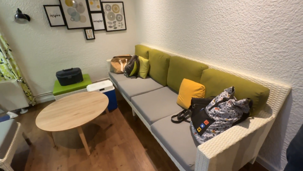 Canapé en osier blanc avec grand coussin rectangle en tissu gris et dossier en tissu vert foncé. Des petits coussins de couleur jaune et vert de chaque côté. Un pouf en osier recouvert de tissu lavable de couleur vert. Table basse en pin de forme ronde. Le mur du cottage premium et blanc avec accroché au mur un ensemble de cadre contour noir et une applique noire.